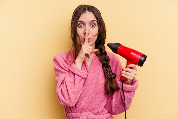 Young woman holding a hairdryer isolated on yellow background keeping a secret or asking for silence.