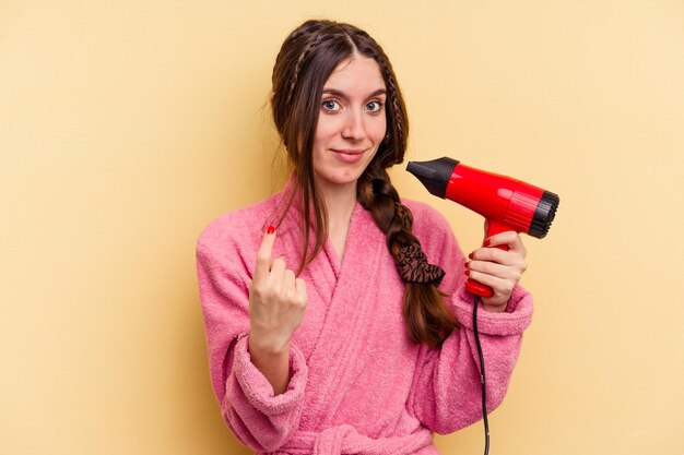 Young woman holding a hairdryer isolated on yellow background pointing with finger at you as if inviting come closer