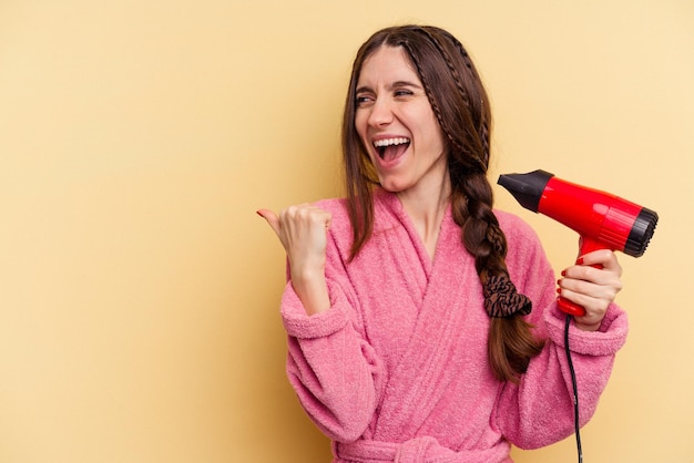 黄色の背景に分離されたヘアドライヤーを持っている若い女性は、親指を離して、笑いながらのんびりとポイントします。