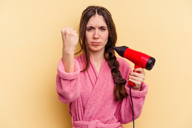 Young woman holding a hairdryer isolated on yellow background showing fist to camera, aggressive facial expression.