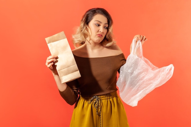 オレンジ色の背景で隔離のプラスチック製の屋内スタジオショットに対してエコロジカルな製品キャンペーンを好む手に紙とビニール袋を持っている若い女性