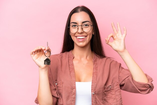 Young woman holding home keys isolated on pink background showing ok sign with fingers