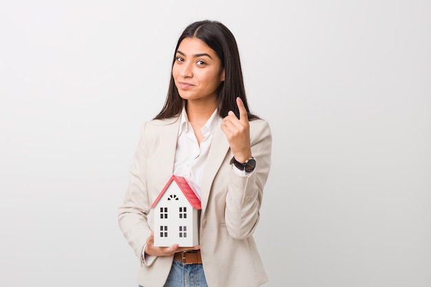 Young woman holding a house icon pointing with finger at you as if inviting come closer