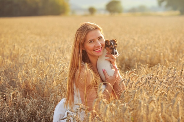 Young woman holding Jack Russell Terrier puppy, in the wheat field, lit by sunset light.