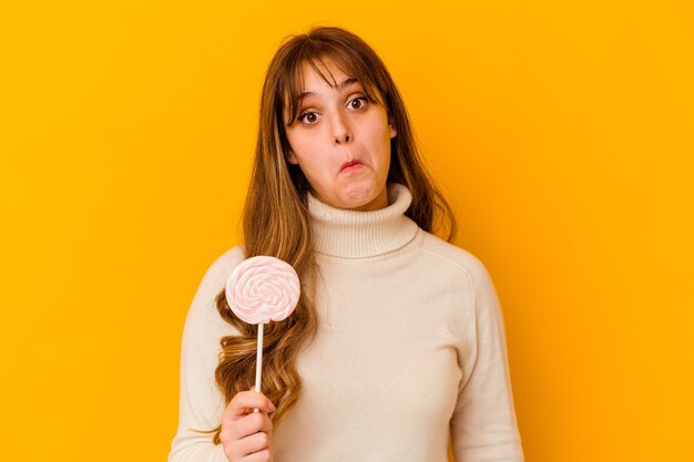 Young woman holding a lollipop isolated on yellow wall shrugs shoulders and open eyes confused