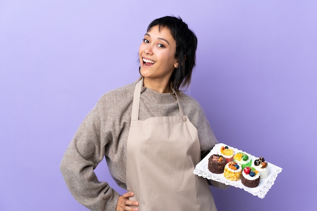 Young woman holding lots of different mini cakes over purple wall applauding