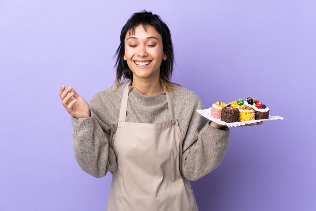 Giovane donna che tiene un sacco di mini torte diverse sul muro viola ridere