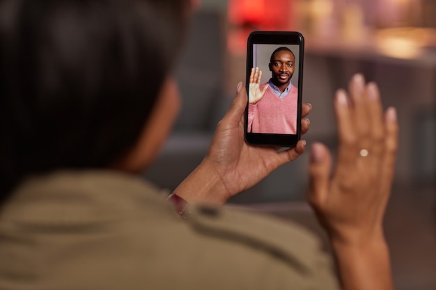 Young woman holding mobile phone and waving to her boyfriend while talking online with him