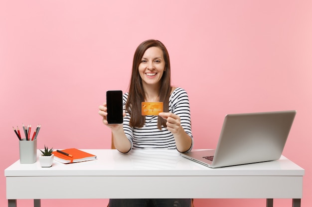 Young woman holding mobile phone with blank empty screen and credit card sit work at white desk with contemporary pc laptop isolated on pastel pink background. Achievement business career. Copy space.