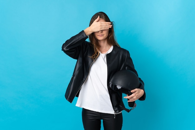 Young woman holding a motorcycle helmet isolated on blue wall covering eyes by hands. Do not want to see something