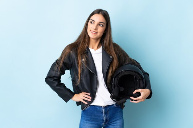 Young woman holding a motorcycle helmet isolated on blue wall posing with arms at hip and smiling