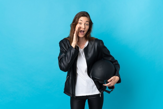 Giovane donna che tiene un casco del motociclo isolato sulla parete blu che grida con la bocca spalancata