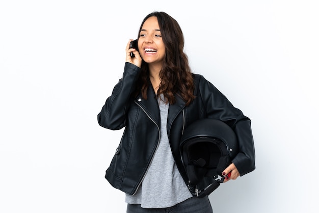 Young woman holding a motorcycle helmet over isolated white wall keeping a conversation with the mobile phone