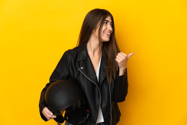 Giovane donna che tiene un casco da motociclista isolato sulla parete gialla che punta di lato per presentare un prodotto