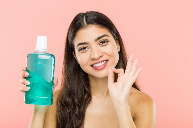 Young woman holding a mouthwash bottle cheerful and confident showing ok gesture.