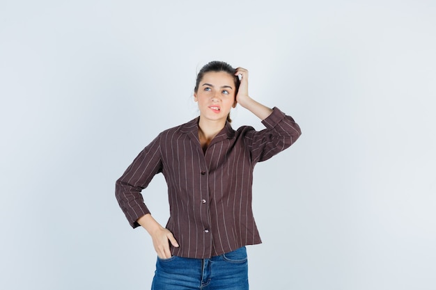 Giovane donna che tiene una mano sulla tempia, un'altra mano sull'anca, morde le labbra in camicia a righe, jeans e sembra dispiaciuta, vista frontale.