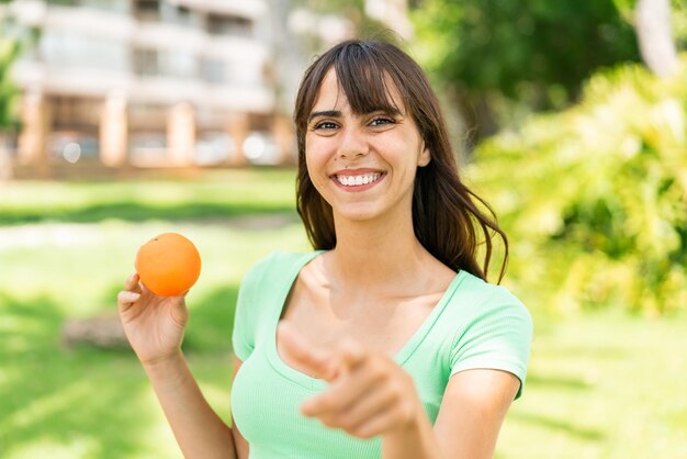 Young woman holding an orange at outdoors points finger at you with a confident expression