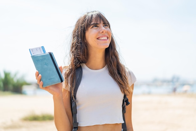 Young woman holding a passport at outdoors looking up while smiling