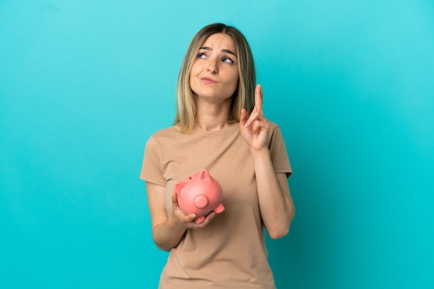 Young woman holding a piggybank over isolated blue background with fingers crossing and wishing the best