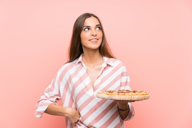 Young woman holding a pizza over isolated pink wall looking up while smiling