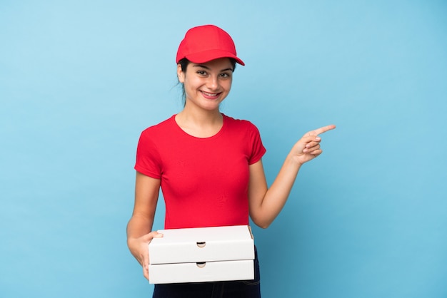 Young woman holding a pizza over isolated pink wall pointing finger to the side