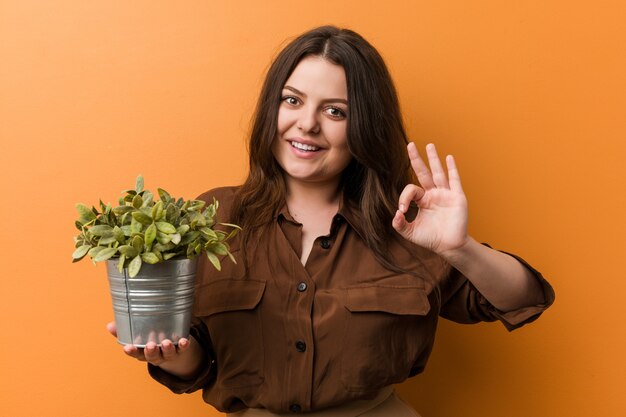 Young woman holding a plant cheerful and confident showing ok gesture.