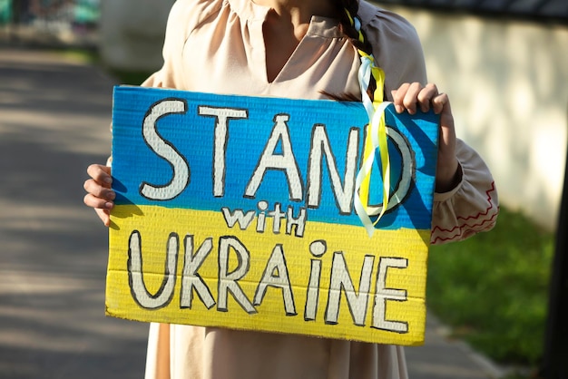 Photo young woman holding poster in colors of national flag and words stand with ukraine outdoors closeup