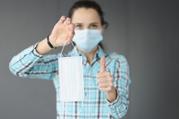 Young woman holding protective medical mask in her hands and showing thumbs up. Mask compliance as prevention of covid 19 pandemic concept