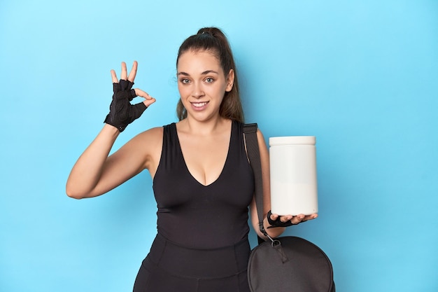 Young woman holding protein bottle in sporty setting cheerful and confident showing ok gesture