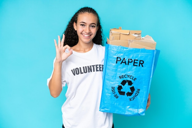 Молодая женщина, держащая мешок для переработки, полный бумаги для переработки, изолирована на синем фоне, показывая пальцами знак "ок"