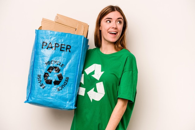 Молодая женщина, держащая мешок для переработки, полный бумаги для переработки на белом фоне, смотрит в сторону, улыбаясь весело и приятно