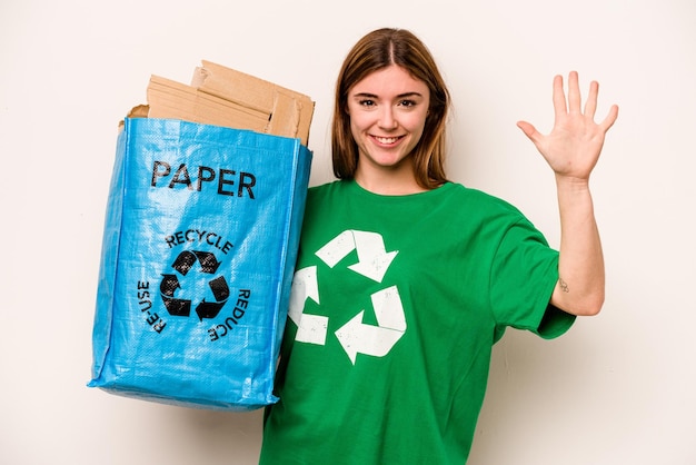 Молодая женщина, держащая мешок, полный бумаги для переработки, изолирована на белом фоне, весело улыбаясь, показывая пальцами номер пять