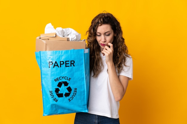 Young woman holding a recycling bag full of paper to recycle isolated on yellow background having doubts