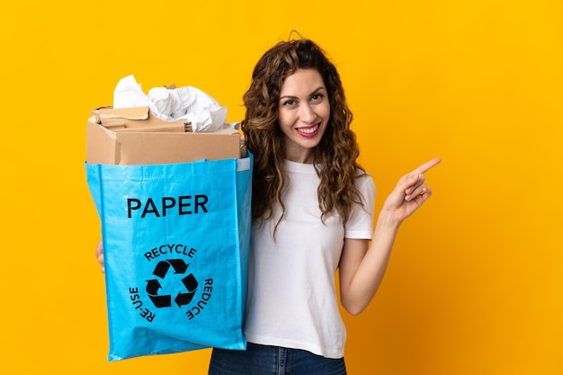 Young woman holding a recycling bag full of paper to recycle isolated on yellow background pointing finger to the side