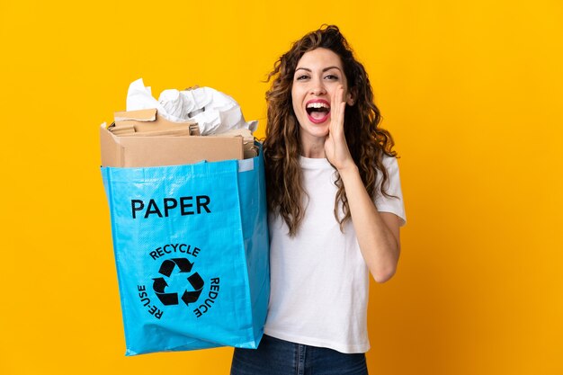 Young woman holding a recycling bag full of paper to recycle isolated on yellow wall shouting with mouth wide open