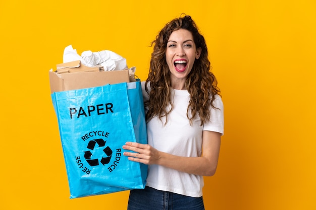 Young woman holding a recycling bag full of paper to recycle isolated on yellow with surprise facial expression