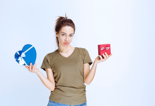 Foto giovane donna che tiene in mano scatole regalo a forma di cuore rosso e blu e sembra confusa a causa della scelta