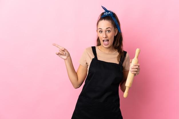 Young woman holding a rolling pin surprised and pointing finger to the side