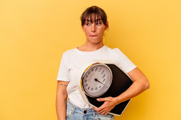 Young woman holding a scale isolated on yellow background confused, feels doubtful and unsure.