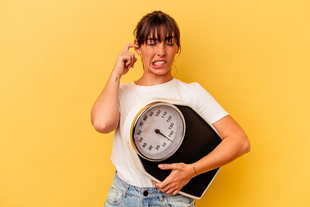 Young woman holding a scale isolated on yellow background covering ears with hands.