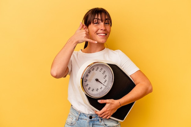 Young woman holding a scale isolated on yellow background showing a mobile phone call gesture with fingers.