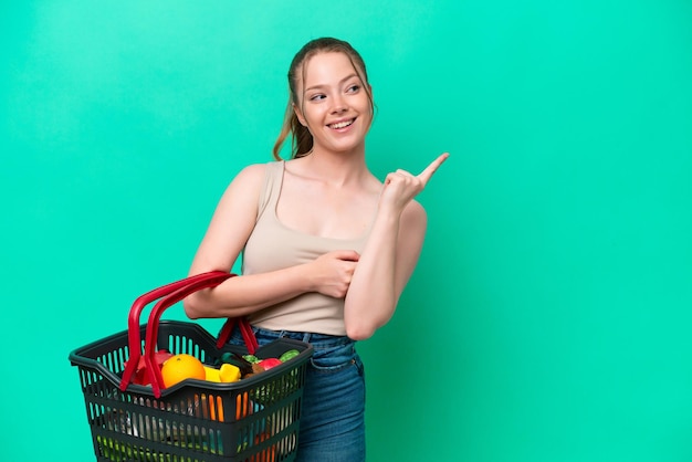 緑の背景に食べ物がいっぱい入った買い物かごを持つ若い女性は、指を上げながら解決策を実現しようとしています