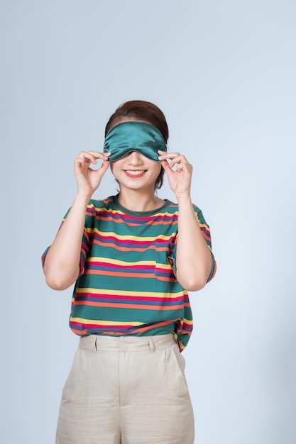 Young woman holding sleeping mask on forehead and looking from under it having fun on pyjama party