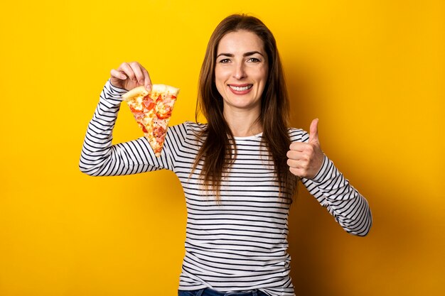 Young woman holding a slice of fresh pizza, showing thumb up on yellow