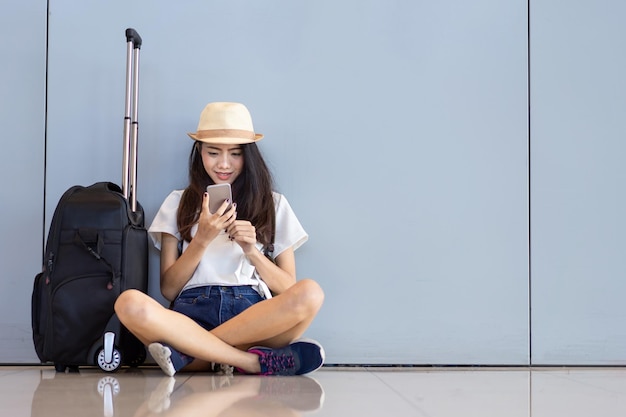 Photo young woman holding smart phone while sitting by luggage on floor