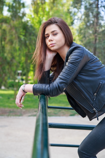 Young woman holding smartphone leaning on fence in park.  