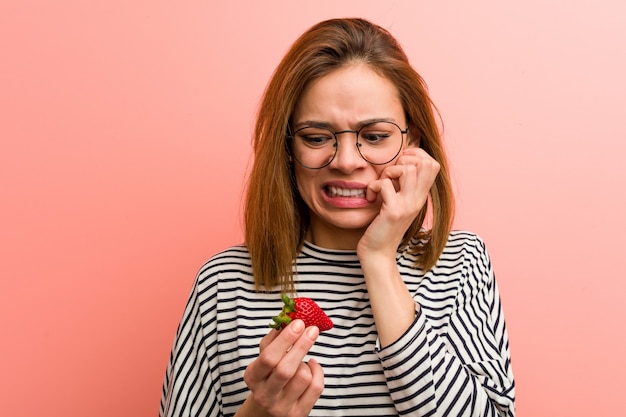 Young woman holding a strawberry biting fingernails, nervous and very anxious.