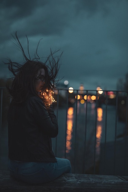 Photo young woman holding string light while sitting on retaining wall over river against sky at dusk