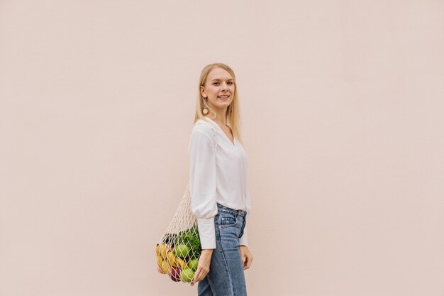 Young woman holding string shopping bag with fruits on a beige background.