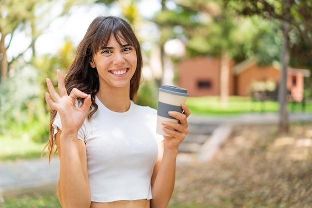 Young woman holding a take away coffee at outdoors showing ok sign with fingers